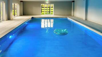 An indoor swimming pool with clear blue water, a green inflatable float, and a view of a green lawn through glass doors.