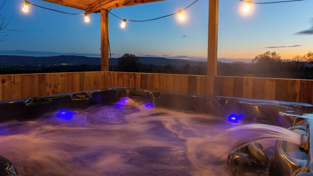 A wooden deck with hot tubs and string lights, overlooking a landscape.