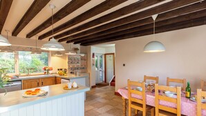 A kitchen with a central island, wooden beams, pendant lighting, and a dining area with a pink tablecloth.