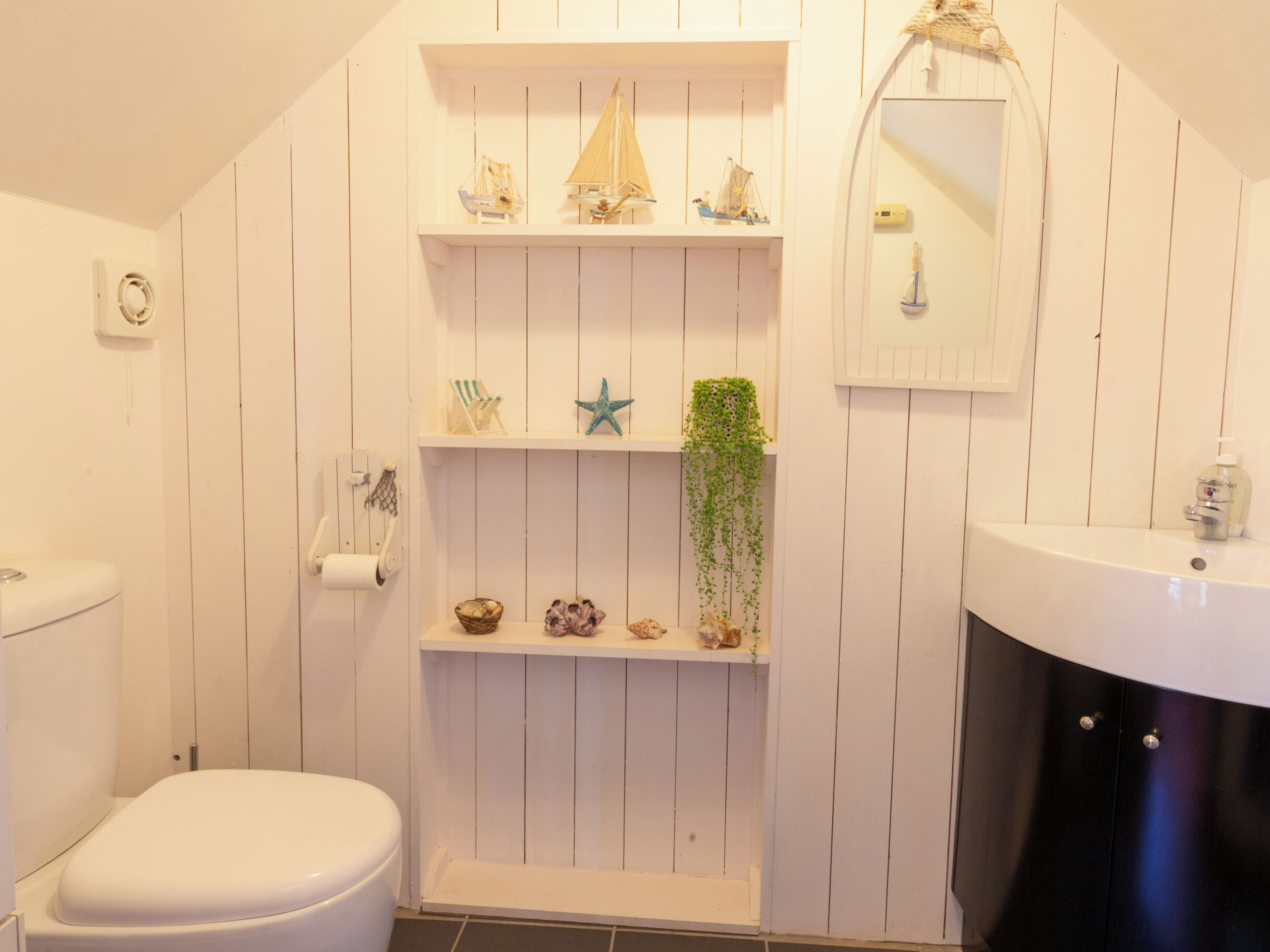 A bathroom with a toilet, a sink, a shelf with decorative items, and a hanging plant.