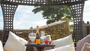 Un área de estar al aire libre con muebles de mimbre, un fondo de pared de piedra, una botella de champán y fresas sobre una mesa pequeña.