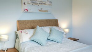 Un lit bien fait, avec une couette blanche à texture, deux oreillers et une tête de lit. On y trouve deux tables de chevet avec des lampes, et un tableau accroché au mur au-dessus du lit.