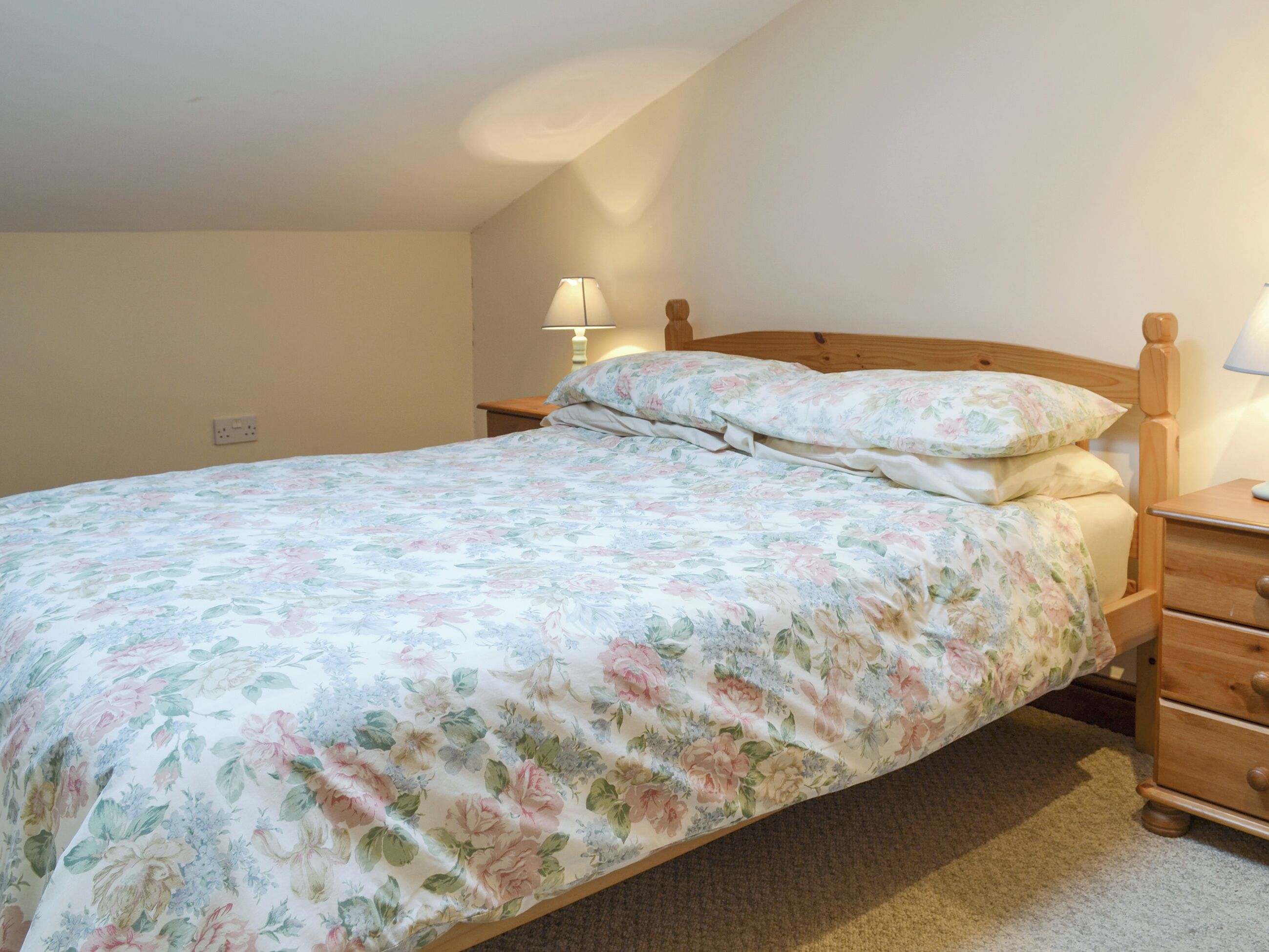 A bedroom with a floral bedspread, a wooden headboard, a nightstand with a lamp, and a dresser.