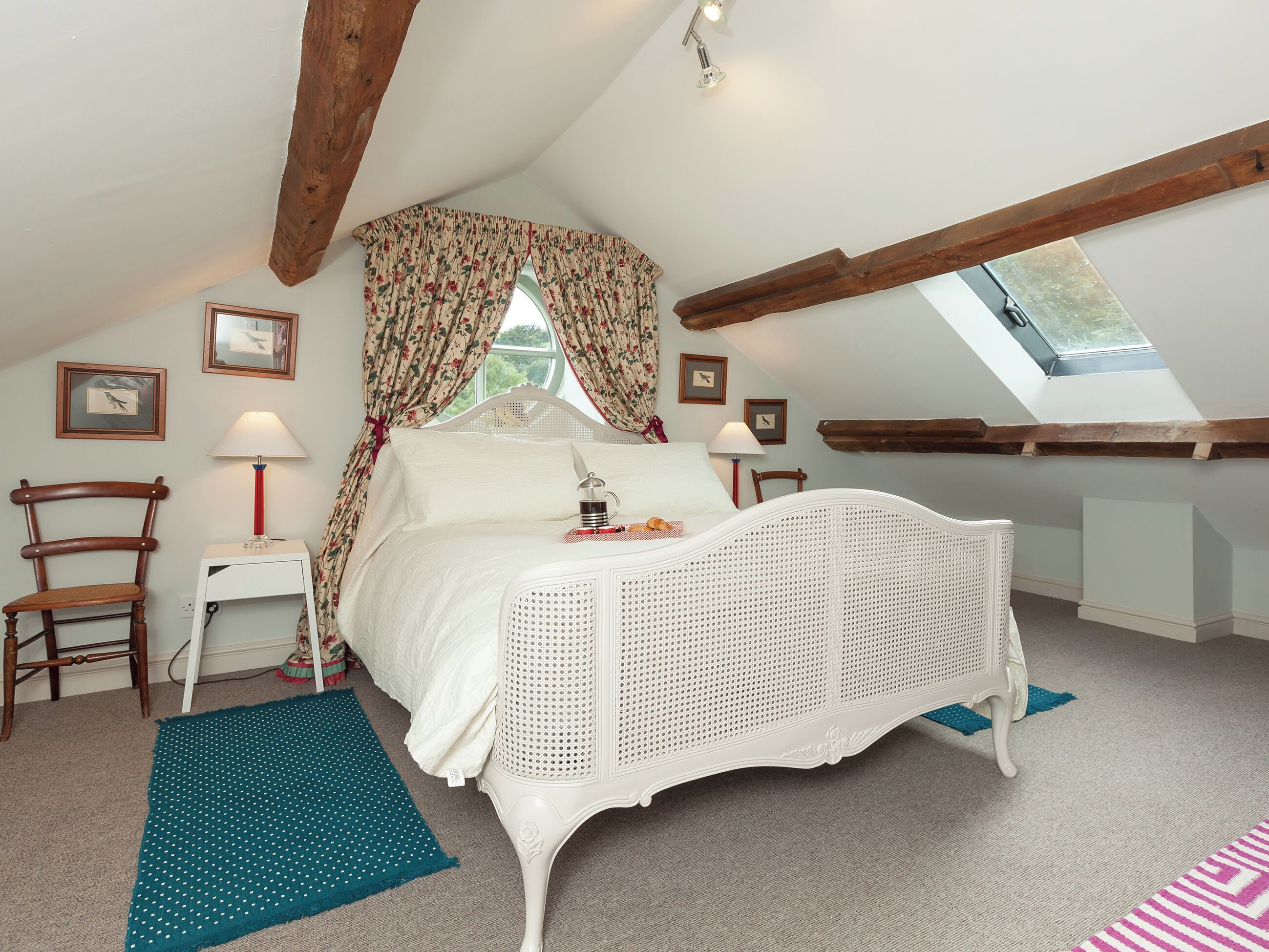 A bedroom with a white bed, floral curtains, a wooden chair, a small bedside table, and a skylight.