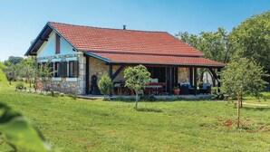 Une maison à deux étages, avec un toit recouvert de tuiles rouges et des murs en pierre, entourée d’une pelouse bien entretenue et d’arbres.