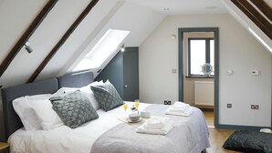 Un dormitorio con techo inclinado, cama con ropa de cama blanca y estampada, una bandeja de desayuno y vistas a un baño a través de una puerta abierta.
