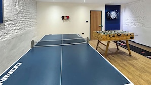 Una mesa de ping pong con el logo 'DONNAY', una mesa de fútbol de mesa y un dardo en una habitación con paredes blancas con textura y suelo de madera.