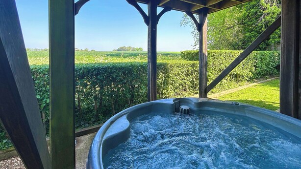 Un jacuzzi bajo una pérgola de madera, con vista a un campo verde y árboles.