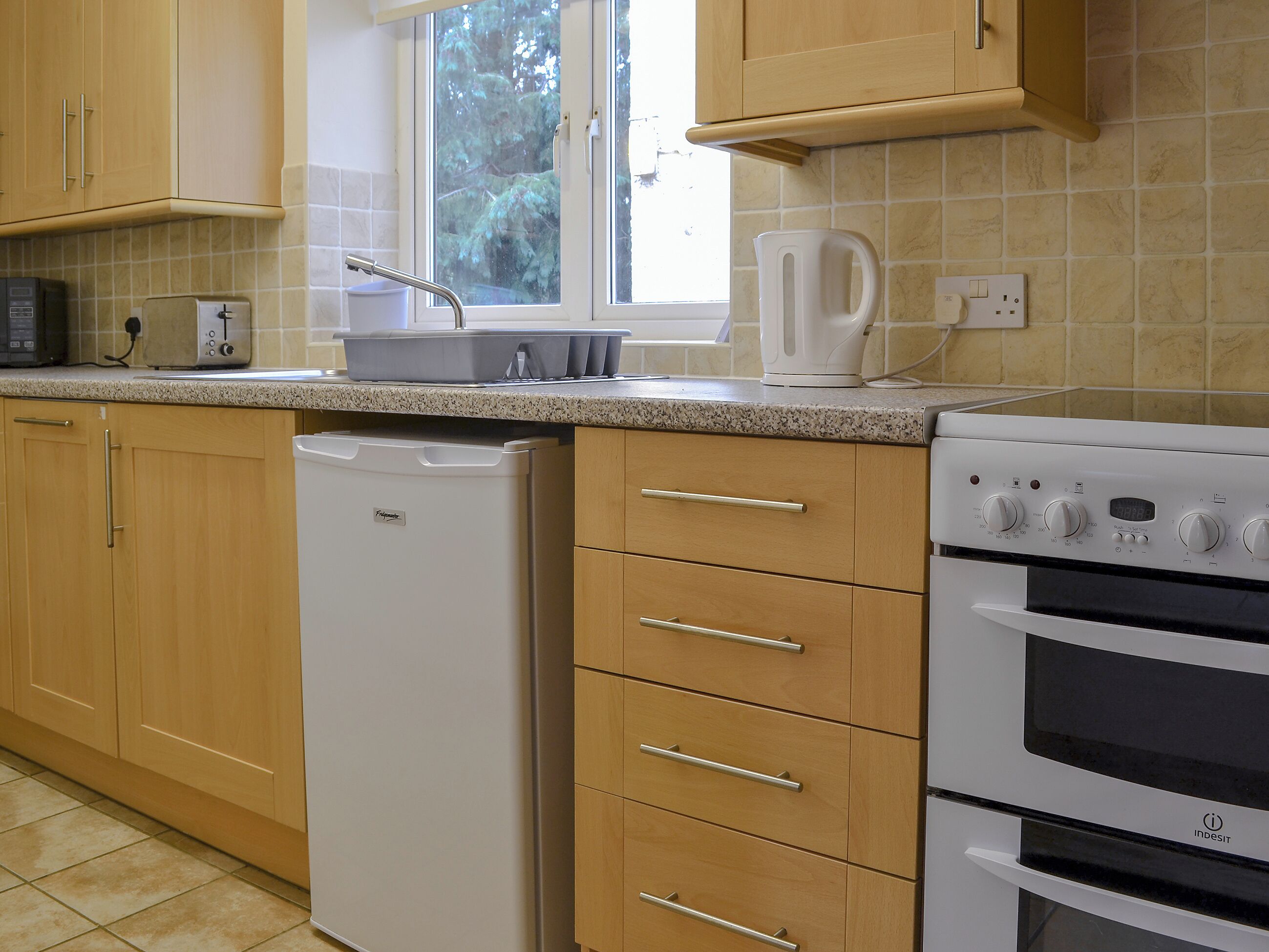 A kitchen with wooden cabinets, a white dishwasher, a stove, and a window with a view of trees.
