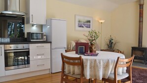 Une cuisine compacte avec des meubles blancs, un four à micro-ondes, un réfrigérateur et un poêle à bois. On y trouve une table à manger avec des chaises et un vase de fleurs.