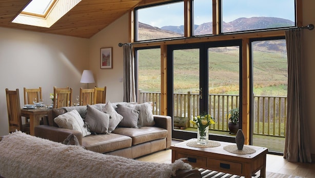 Een gezellige woonkamer met een houten plafond, een bank, een eettafel met stoelen en een groot raam met uitzicht op het landschap.