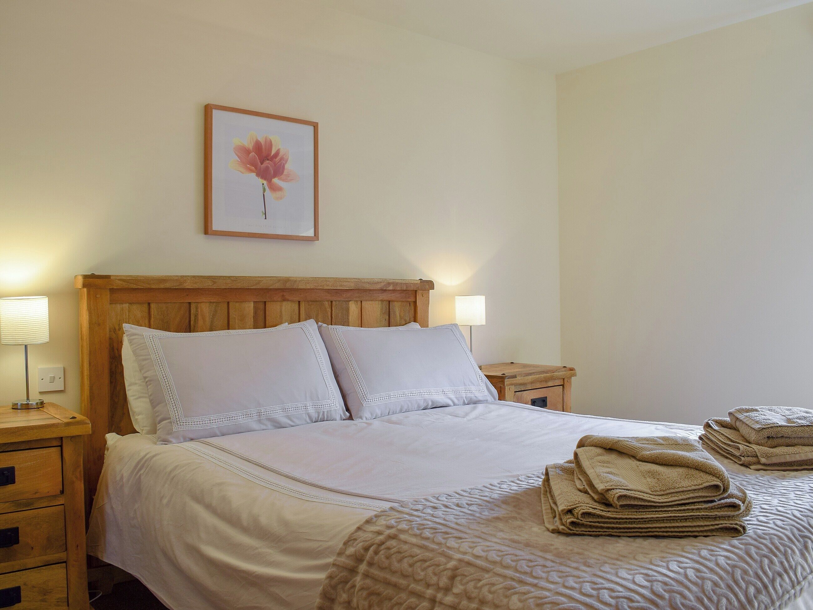 Un lit bien fait, avec du linge de lit blanc et un plaid beige, une tête de lit en bois, deux tables de chevet avec des lampes et un tableau encadré au mur.