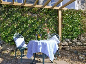An outdoor table set with a checkered tablecloth, two wine glasses, a bottle, and a vase with flowers, under a wooden pergola with a stone wall backdrop. - 4 bedroom accommodation in Llanaber, near Barmouth (Llanaber, near Barmouth)