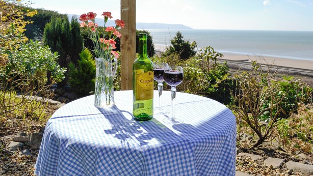 Una mesa con una botella y vasos, un florero con flores y vista a la playa.