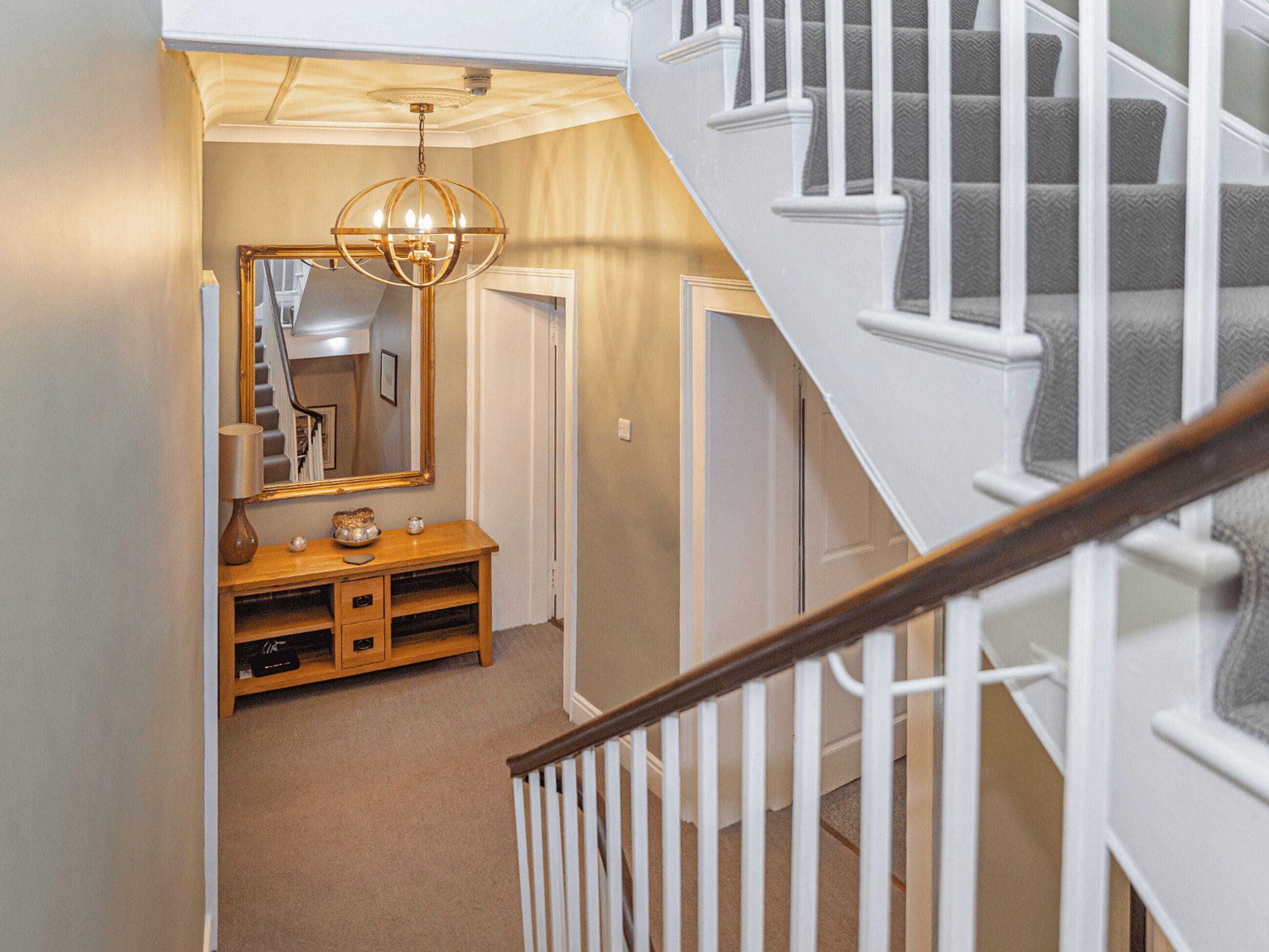 A staircase with white railings and a wooden handrail, leading to a landing with a mirror, a lamp, and a small table.