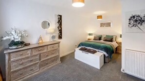 Une chambre avec une commode en bois, un lit avec une literie verte, une table de chevet blanche et un radiateur.