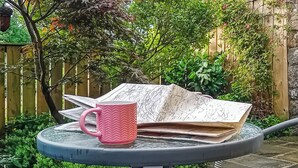 Ein runder Tisch mit einer rosa Tasse und einer Karte, umgeben von Grünpflanzen und einem Holzzaun.