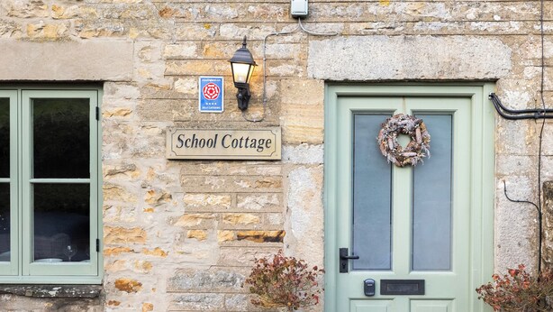 Ein Steinhaus mit einer grünen Tür, einem Schild mit der Aufschrift 'School Cottage', einem Laternen und einem Kranz.