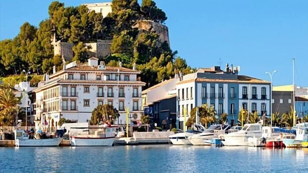 Una marina con barcos amarrados, edificios con balcones y un acantilado con árboles al fondo.