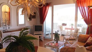 Amplia sala de estar con televisor, mobiliario de mimbre blanco, una planta grande y un balcón con asientos exteriores.