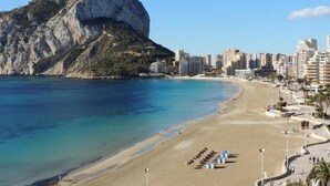Un paisaje costero con una montaña rocosa destacada, una playa arenosa y una hilera de modernos edificios altos.