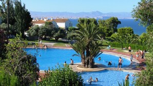 Zona de piscina con gente nadando y tomando el sol, rodeada de exuberante vegetación y vistas al mar y montañas a lo lejos.