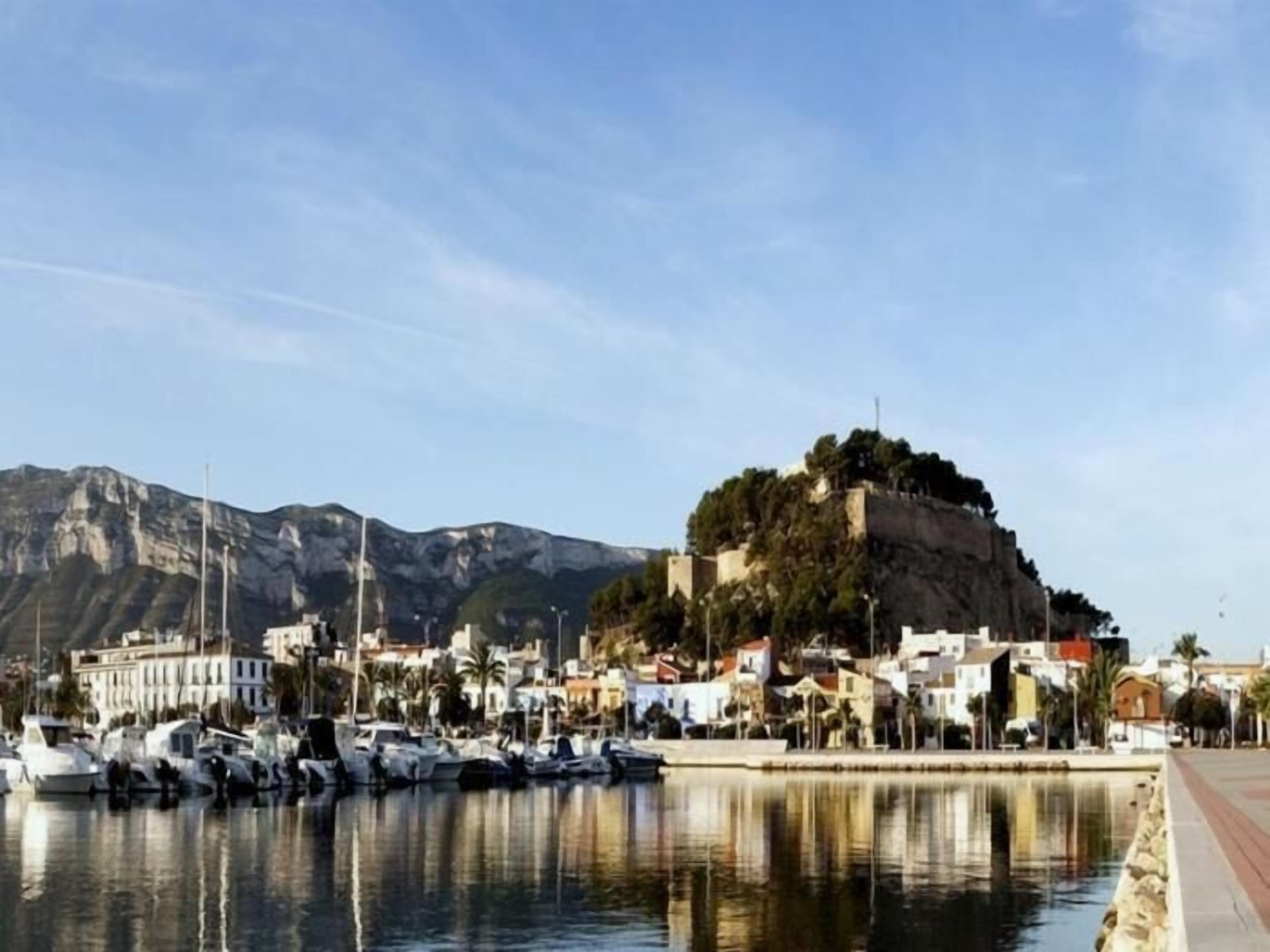 A coastal marina with boats docked, a prominent hill with buildings, and a clear sky.