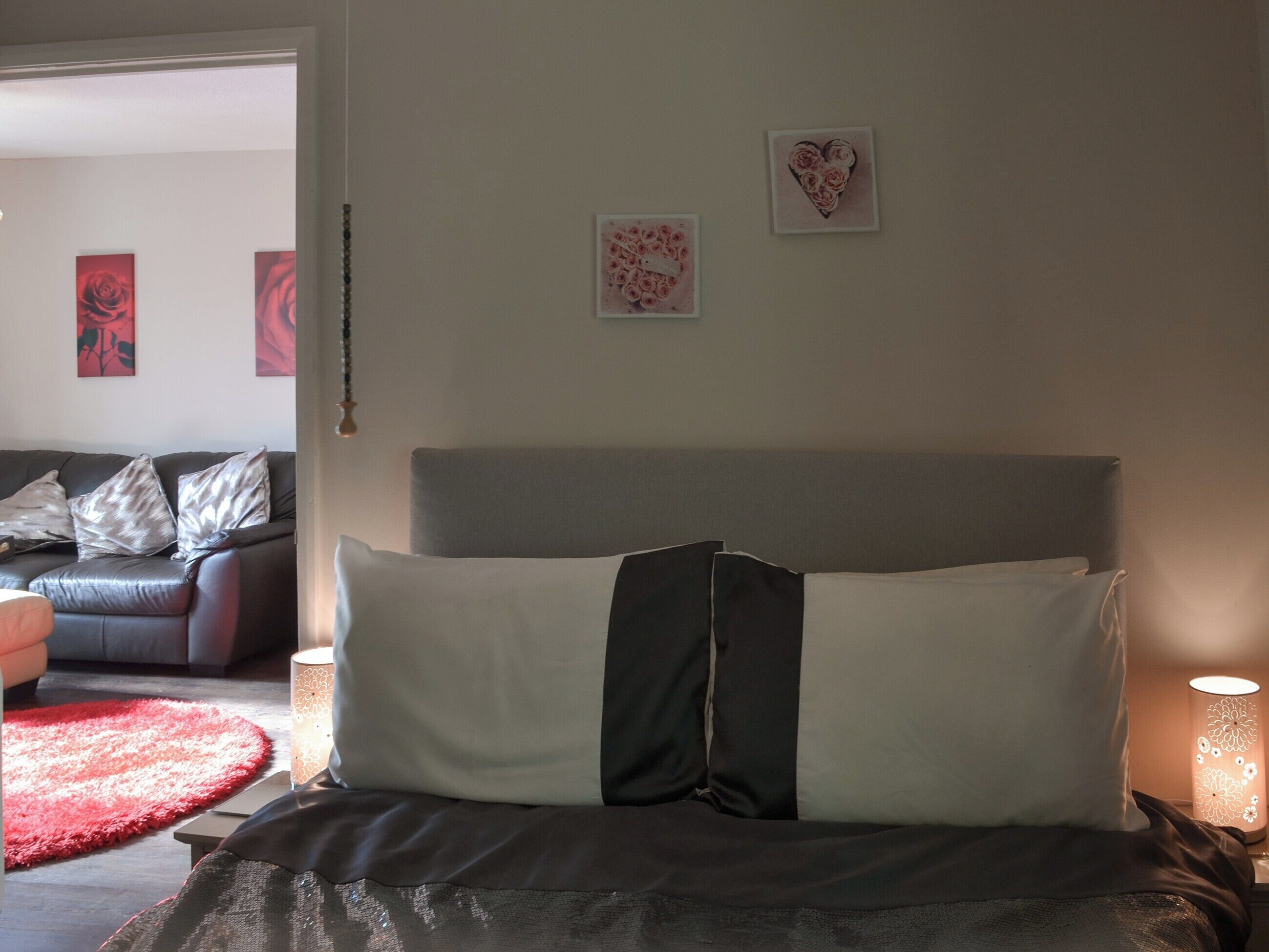 A bedroom with a bed, a nightstand, a lamp, a red rug, and a couch in the background.