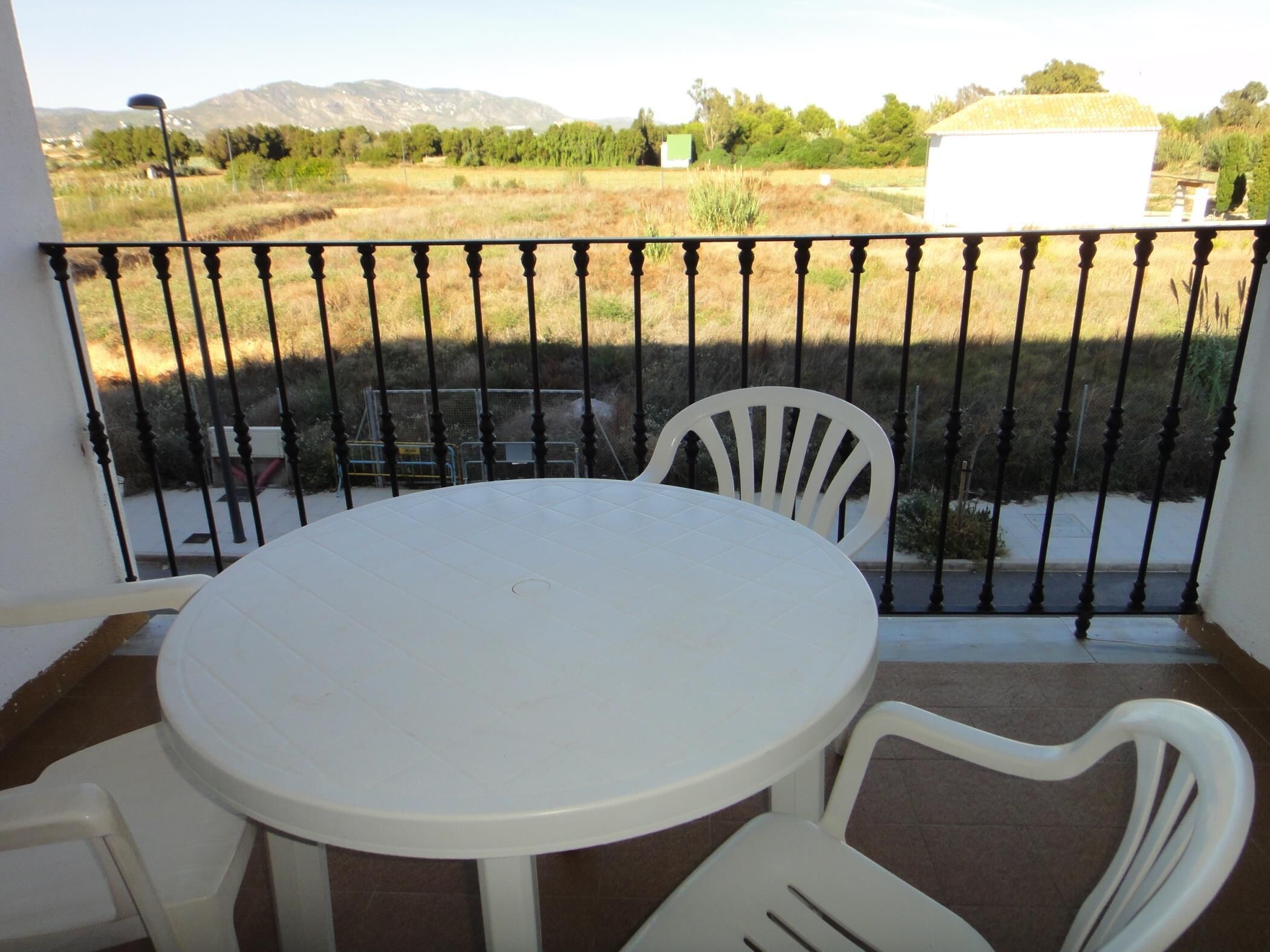 A balcony with a round table and two chairs, overlooking a rural landscape with fields and distant mountains.