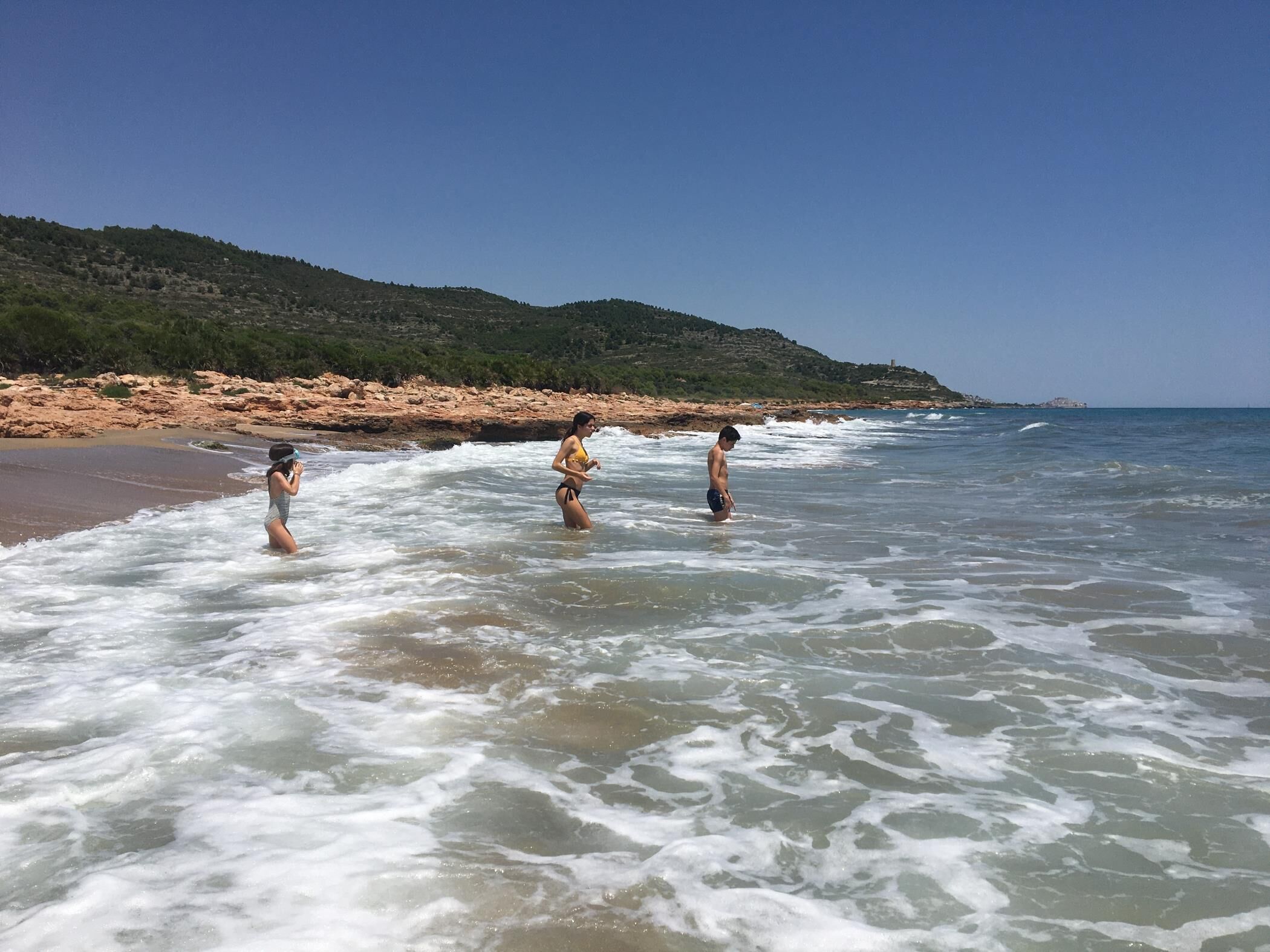 Ein Strand mit drei Personen im Wasser, einer felsigen Küste und einem klaren Himmel.