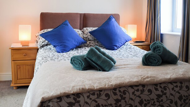 Una cama bien hecha con una colcha estampada, dos almohadas azules y dos toallas enrolladas sobre la cama.