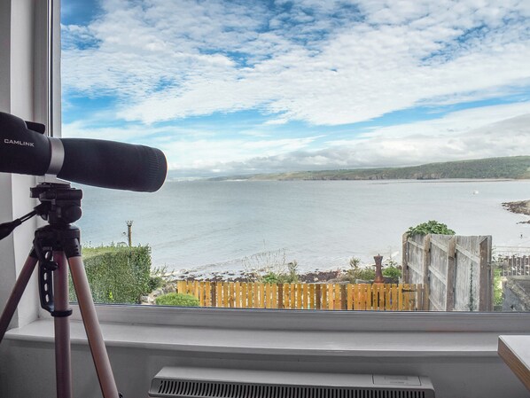 A telescope on a tripod by a window overlooking a body of water with a wooden fence in the foreground.