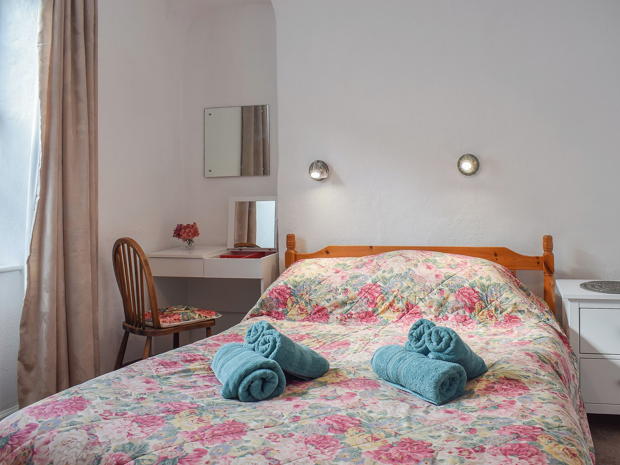Un dormitorio con colcha estampada, cabecera de madera, mesita de noche, una silla, escritorio con espejo y un florero con flores.