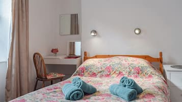 Un dormitorio con colcha estampada, cabecera de madera, mesita de noche, una silla, escritorio con espejo y un florero con flores.