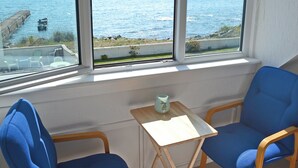 Une pièce avec vue sur la mer, deux chaises bleues, une petite table et une fenêtre donnant sur une côte rocheuse.