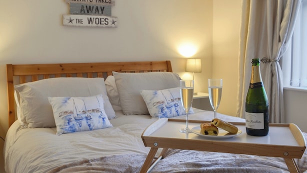 Een netjes opgemaakt bed met kussens, een nachtkastje met glazen en een fles champagne, en een bordje aan de muur.