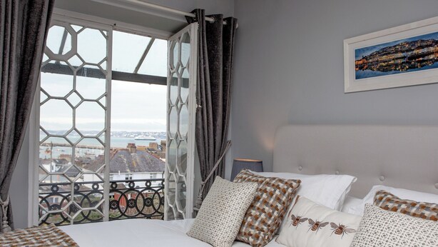 A bedroom with a large bed, a view of a cityscape through a window with a metal railing, and a framed picture on the wall. - 8 bedroom accommodation in Brixham (Brixham)