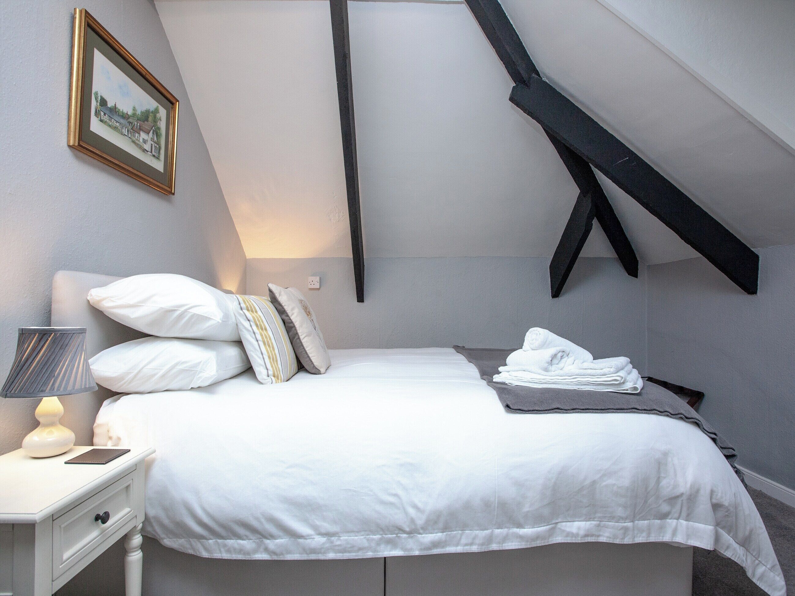 A bedroom with a bed, pillows, a bedside lamp, a folded towel, and a framed picture on the wall.
