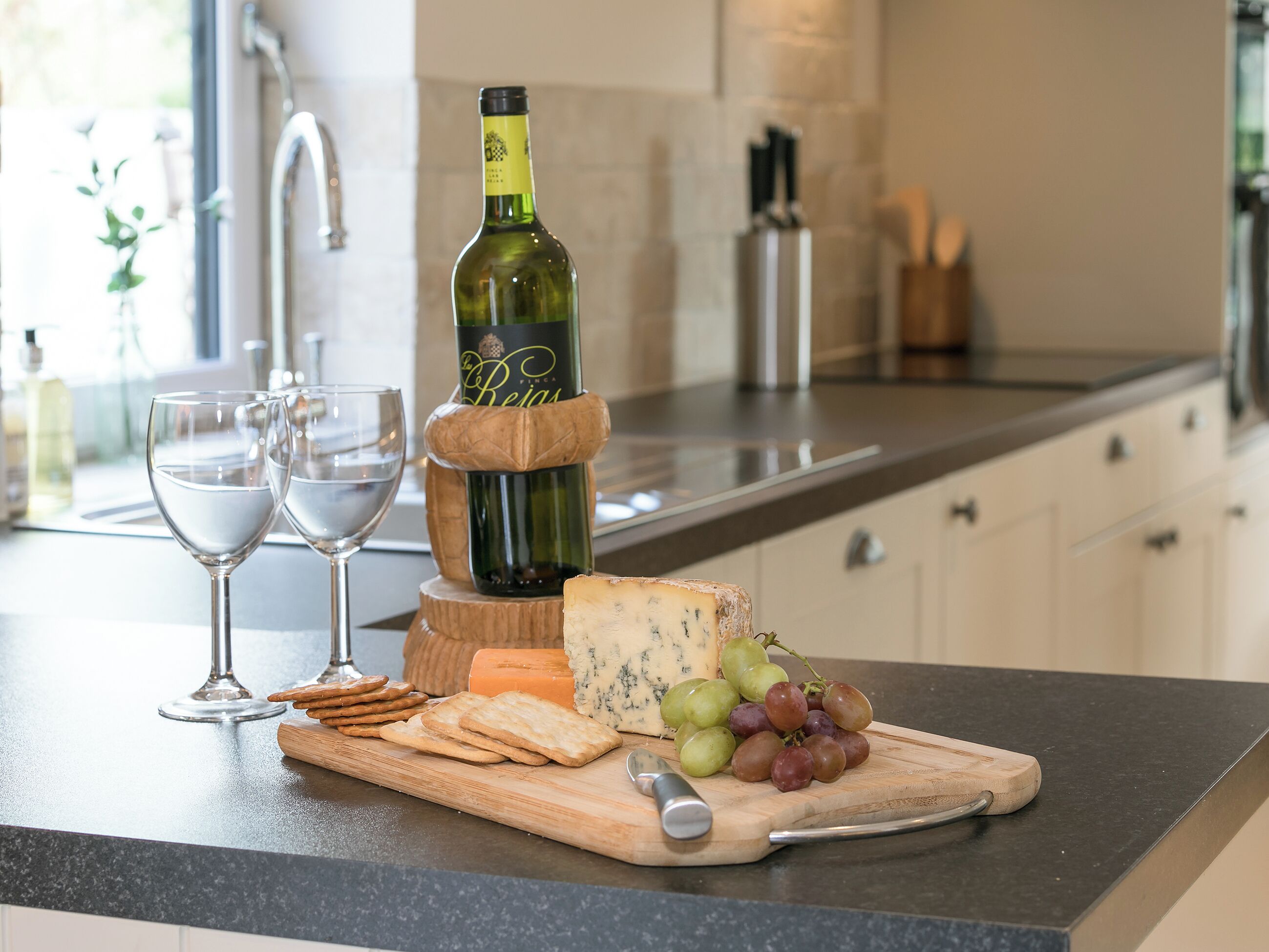 Una encimera de cocina con una botella de vino, dos copas, queso, galletas y uvas.