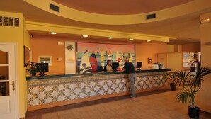 Un hall d'accueil d'hôtel avec un sol carrelé, un comptoir de réception, une grande fresque murale et des plantes en pot.