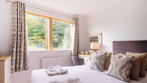A bedroom with a bed, a nightstand, a lamp, a window with curtains, and a view of greenery outside. - North Bay Sands Apartment 2- UK2630 (Scarborough)