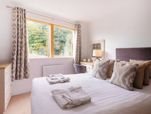 A bedroom with a bed, a nightstand, a lamp, a window with curtains, and a view of greenery outside.