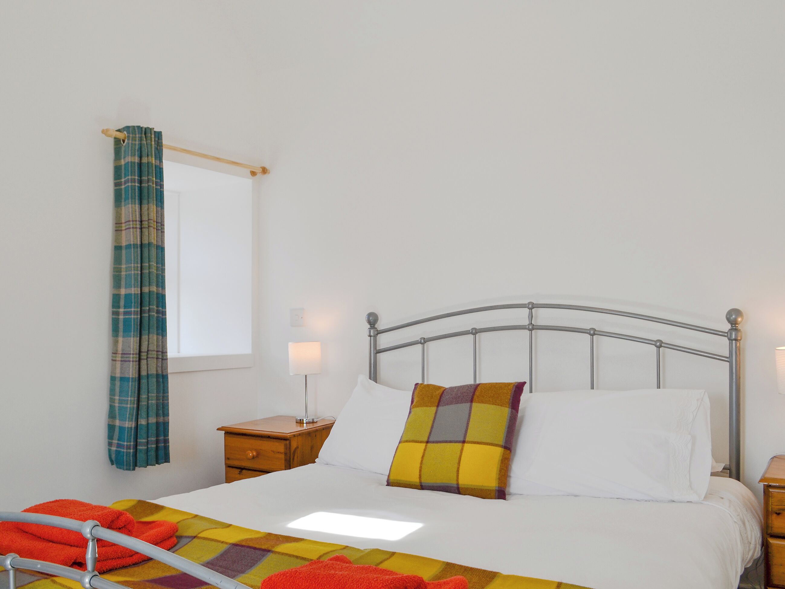 A neatly made bed with white linens and a colorful throw blanket, a metal-framed headboard, a wooden nightstand with a lamp, and a window with a plaid curtain.