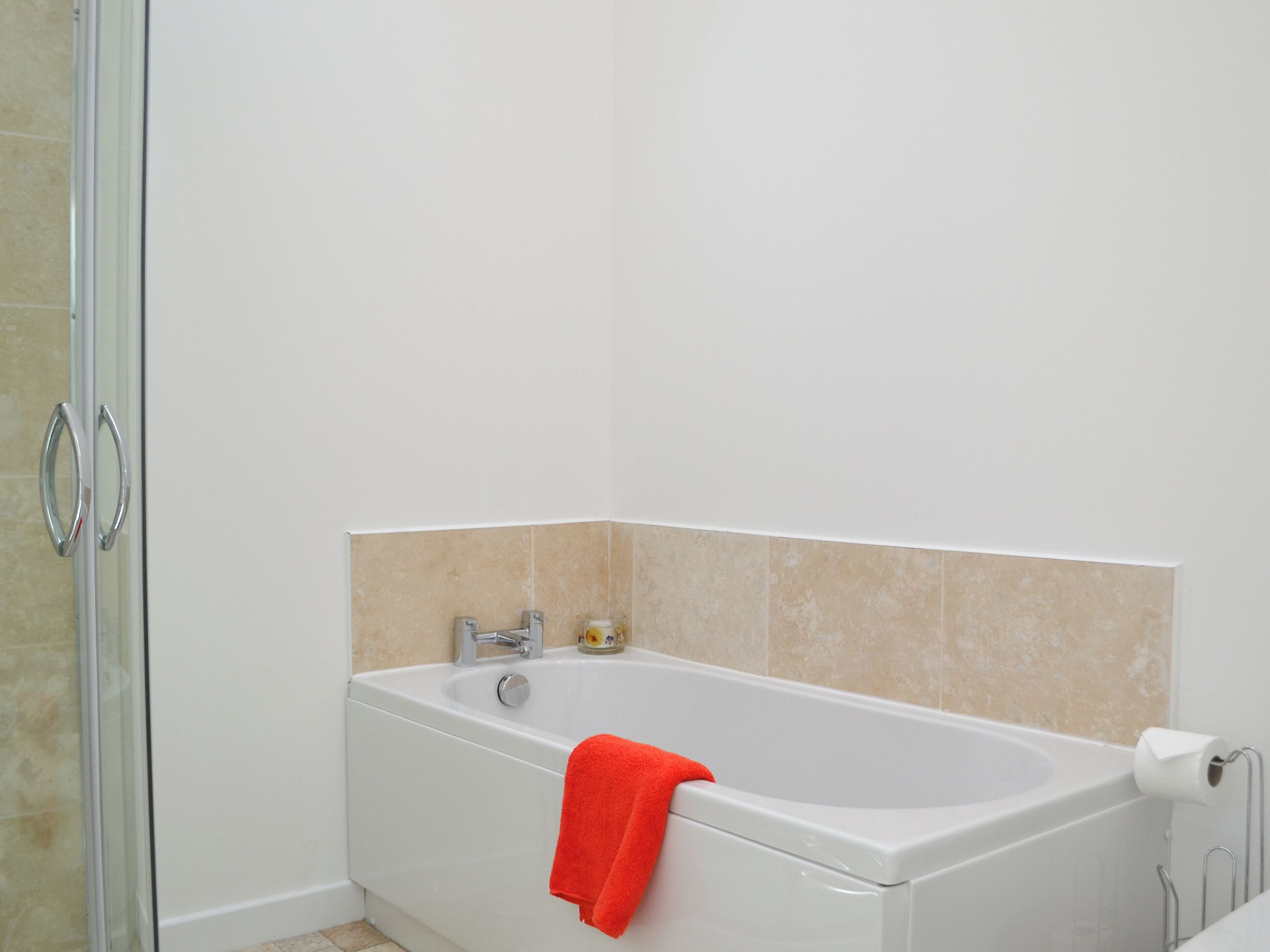 Un bagno con una vasca da bagno bianca, box doccia in vetro, piastrelle color beige e un asciugamano rosso.