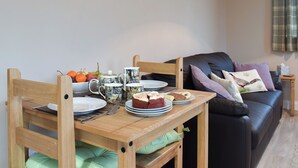 Une table à manger avec des assiettes, une théière et un saladier rempli de fruits. Il y a un canapé et un tableau accroché au mur.