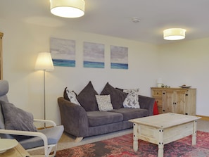 Sala de estar con un sofá gris, un sillón blanco, una mesa de centro de madera y un tapiz rojo estampado. Se observan tres cuadros colgados en la pared, una lámpara de pie y un mueble de madera con un detalle rojo.