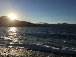 En strand där solljuset reflekteras i vattnet, med berg i bakgrunden och en klar himmel.