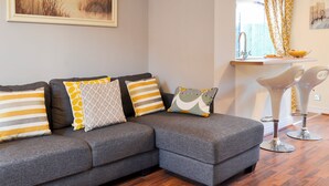 Una sala de estar moderna con un sofá seccional gris, un cuadro de pastos altos y una zona de cocina con una cortina amarilla estampada.