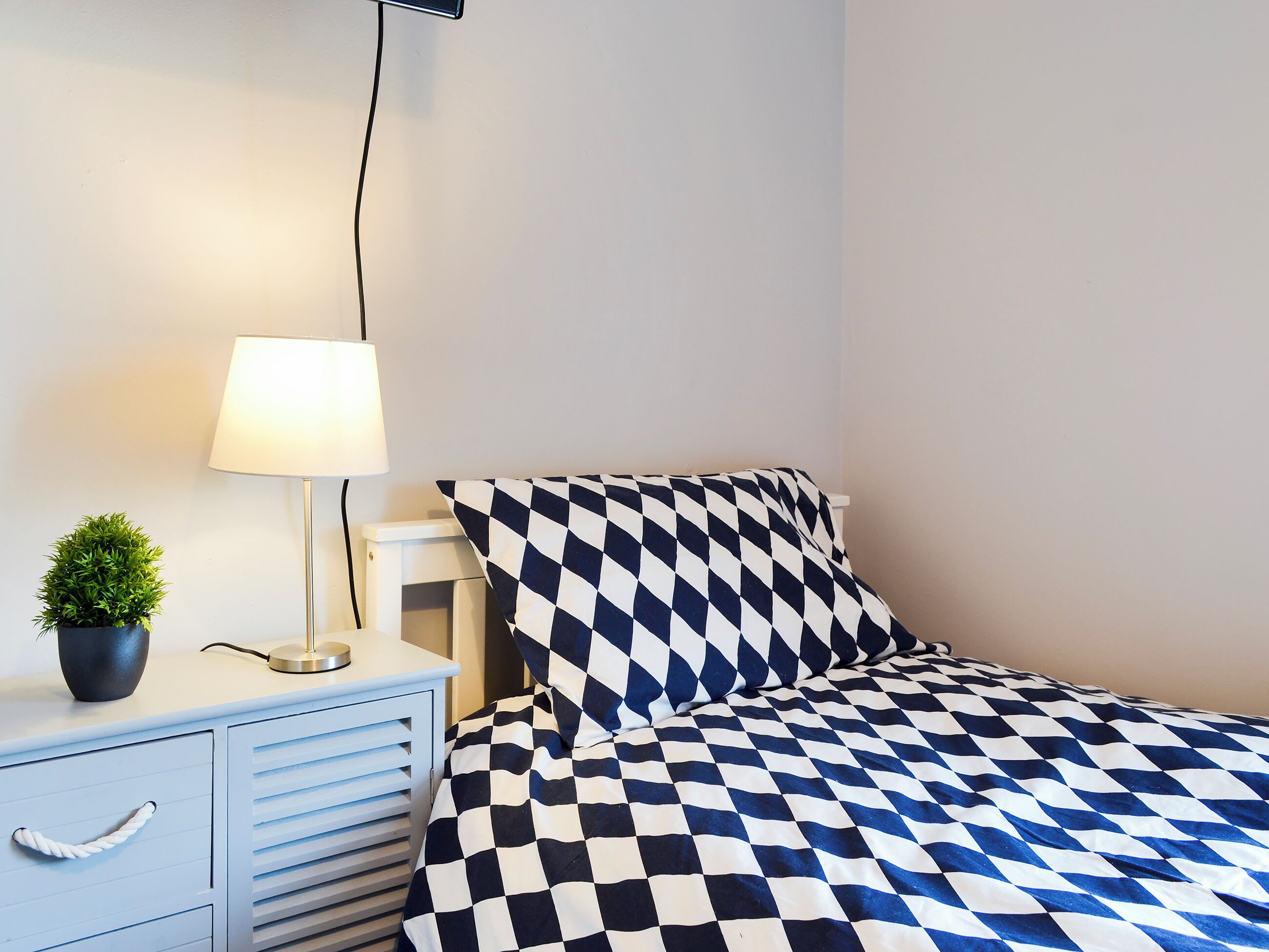 Ein ordentlich eingerichtetes Schlafzimmer mit karierter Bettdecke, einer Nachttischlampe, einem kleinen Topfpflanzen und einem weißen Nachttisch mit Schubladen.