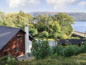 Une cabane avec un toit en pente, une cheminée et une vue sur un lac et des collines boisées.